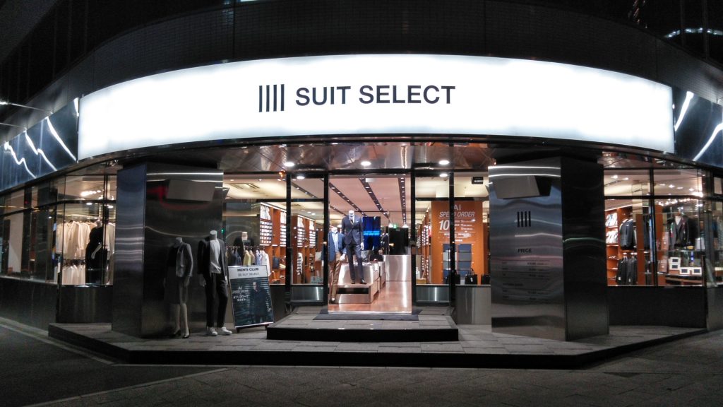 スーツセレクト横浜東店の店頭画像。ガラス張り正面にはスーツスタイルのマネキンが見える。店頭には数体のマネキン。夜間の画像なので、店内の照明でとても明るい店頭。